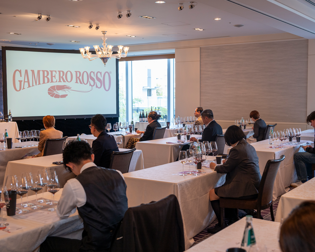 Group of sommeliers sitting at their desks in a wine tasting room in front of a big screen with red Gambero Rosso logo on a white background