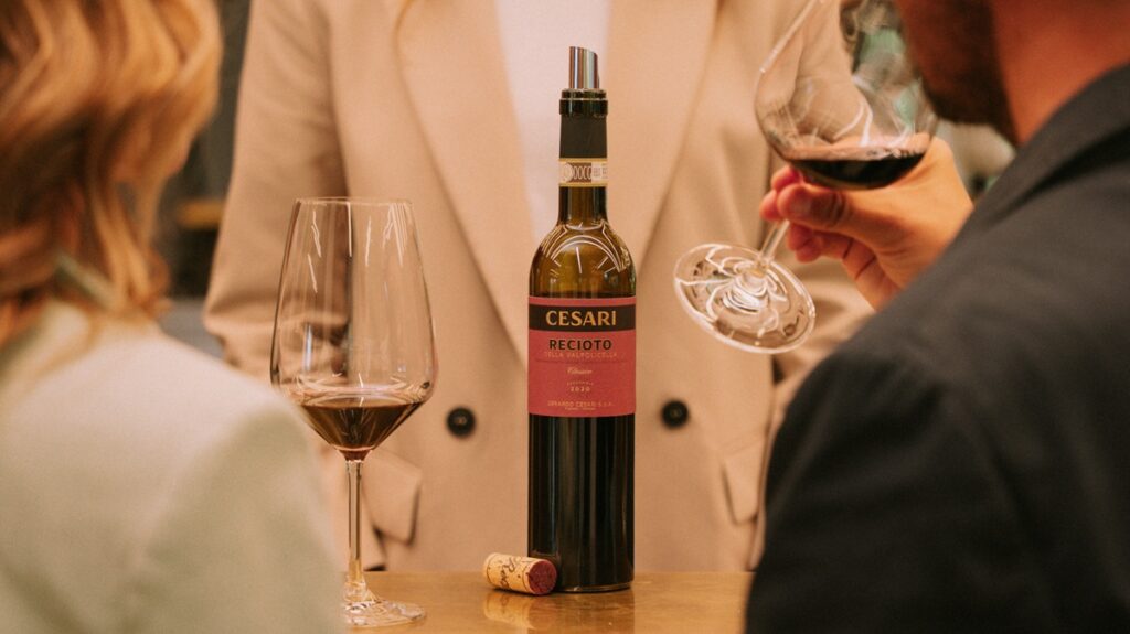 Recioto della Valpolicella Cesari bottle standing on a desk with a person behind it, and two people in the front drinking from wine glasses