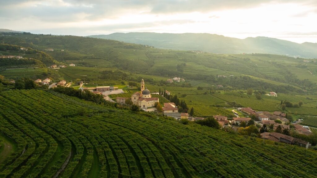 Wine Tour Valpolicella _Aerial view of the historic village of Castelrotto in Valpolicella, featuring stone houses and a church dome nestled among lush green terraced vineyards under a cloudy sky with sunlight breaking through.